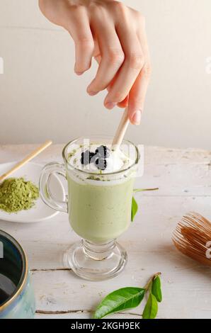 Matcha Latte mit Schlagsahne und Bluebberies, weibliche Hand mit einem Trinkhalm. Stockfoto