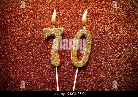 Brennende goldene Kerze mit der Nummer 70 auf rotem glitzernden Hintergrund Stockfoto