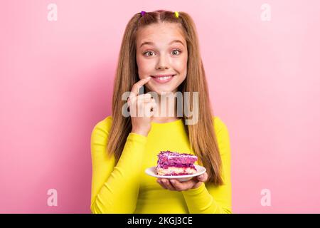 Foto eines gerissenen, trickreichen Schulmädchens mit gelben Fingerlippen und einem Mund, der Kuchenstückchen hält, isolierter rosafarbener Hintergrund Stockfoto