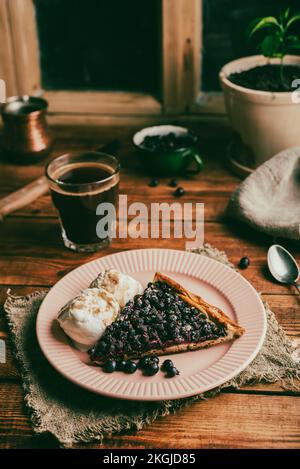 Ein Stück frische hausgemachte Amelanchier-Beeren-Galette und zwei Kugeln Vanille-Eiscreme auf dem Teller mit einem Glas Kaffee auf dem rustikalen Tisch Stockfoto