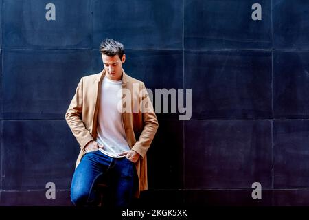 Junger Mann, der Winterkleidung auf der Straße trägt. Junger Kerl mit moderner Frisur mit Mantel, blauer Jeans und weißem Sweeter. Stockfoto