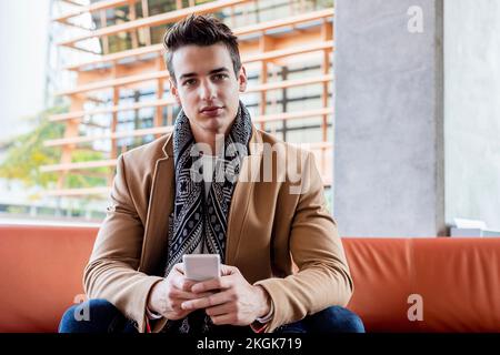 Ein junger erwachsener Kaukasier, der im Herbst draußen auf der Bank sitzt, sein Handy hält und in die Kamera schaut Stockfoto