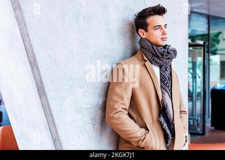 Junger Mann, der Winterkleidung auf der Straße trägt. Junger Kerl mit moderner Frisur mit Mantel, blauer Jeans und weißem Sweeter. Stockfoto