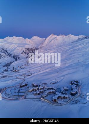 Luftaufnahme der Bergdorf Chalets beleuchtet mit nächtlichen Straßenlaternen entlang Serpentinen kurvige Straße in der Dämmerung bei Sonnenuntergang. Skigebiet mit Schnee Stockfoto