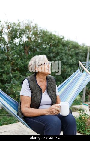 Wunderschöne ältere blonde Frau, die Buch liest und in einer Hängematte im Garten sitzt Stockfoto