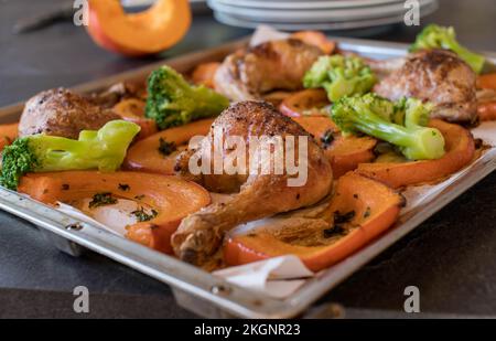 Knusprig gebackene Hähnchenschenkel mit rotem Kuri-Kürbis und Brokkoli auf einem Backblech auf dem Küchentisch. Vorderansicht mit unscharfem Hintergrund Stockfoto