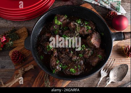 Weihnachtsfleischgericht mit geschmorten Schweinefilet-Medaillons und Soße aus roten Zwiebeln in einer rustikalen Pfanne auf einem Holztisch mit Tellern, Servietten und Ziergegenständen. Stockfoto