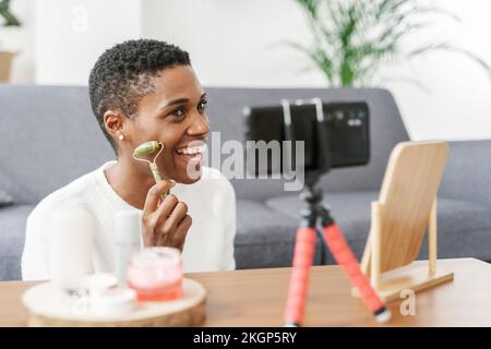 Glückliche Frau, die zu Hause mit ihrem Smartphone mit Jade Roller arbeitet Stockfoto