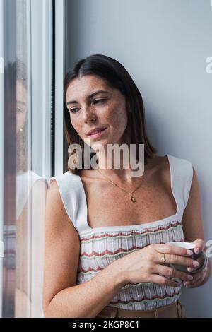 Aufmerksame Geschäftsfrau mit Kaffeetasse, die durch das Fenster im Büro schaut Stockfoto