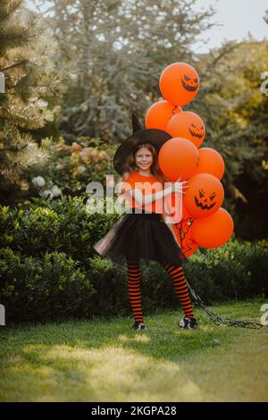 Lächelndes Mädchen mit Hexenhut und orangefarbenen Ballons im Garten Stockfoto