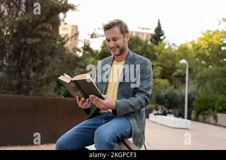 Ein glücklicher Mann, der auf einer Bank im Park ein Buch liest Stockfoto