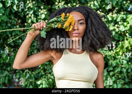 Junge Frau, die das Auge vor der Pflanze mit gelben Blüten bedeckt Stockfoto
