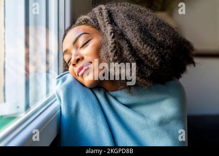 Junge Frau, die zu Hause in eine Decke gewickelt ist und sich an das Fenster lehnt Stockfoto