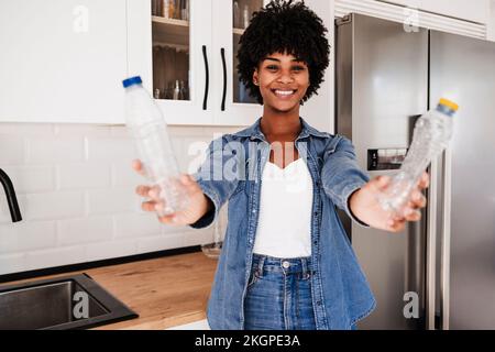 Glückliche junge Frau, die Plastikflaschen zeigt, die zu Hause in der Küche steht Stockfoto