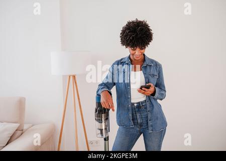 Lächelnde Frau, die mit dem Staubsauger steht und zu Hause ein Smartphone benutzt Stockfoto
