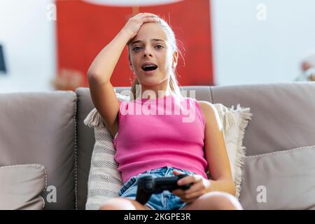 Ein Mädchen mit Kopf in der Hand, das den Joystick hält und zu Hause auf dem Sofa sitzt Stockfoto