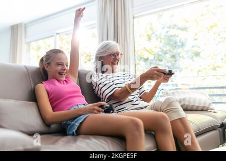 Enkeltochter mit handgehaltener Hand spielt Videospiel, während Großmutter zu Hause auf dem Sofa sitzt Stockfoto