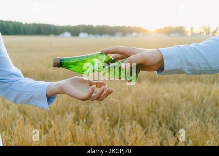 Die Hand eines Mannes, der eine zerbrochene Plastikflasche an eine Frau auf dem Feld weitergibt Stockfoto