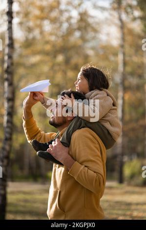 Vater trägt einen Jungen, der im Park ein Papierflugzeug auf den Schultern hält Stockfoto