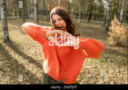 Lächelnde Frau in orangefarbenem Pullover, die sich mit den Händen im Park in Form eines Herzens macht Stockfoto