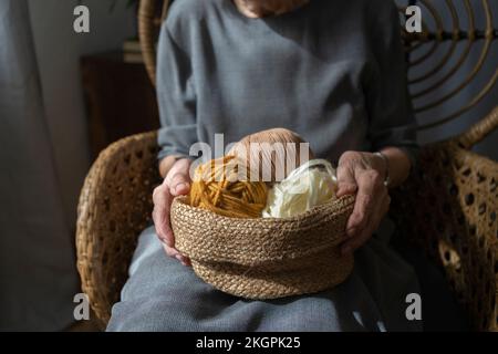 Hände einer älteren Frau, die zu Hause einen Korb aus farbenfroher Strickwolle hält Stockfoto