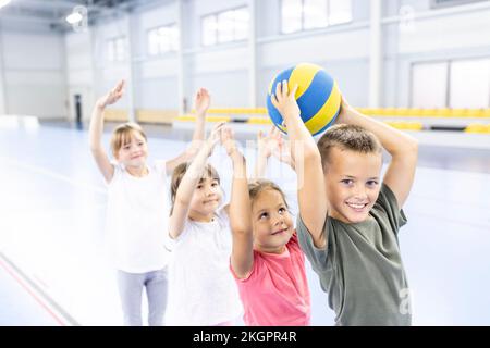 Lächelnder Schuljunge, der den Ball an Freunde in der Schlange auf dem Sportplatz weitergibt Stockfoto