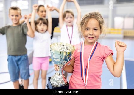 Ein Mädchen mit Medaille und Trophäe feiert den Sieg mit Freunden auf dem Schulsportplatz Stockfoto