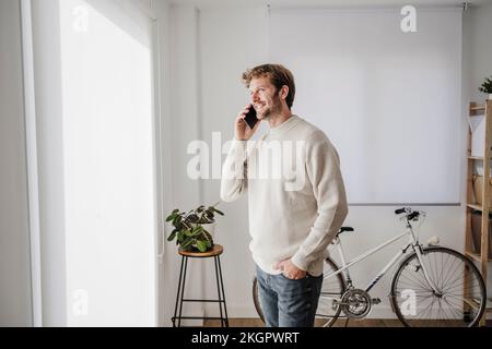 Glücklicher Geschäftsmann, der mit dem Smartphone spricht und aus dem Fenster am Arbeitsplatz schaut Stockfoto