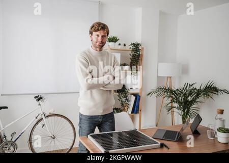 Selbstbewusster Geschäftsmann, der die Arme von einem Solarmodul auf dem Schreibtisch gekreuzt hat Stockfoto