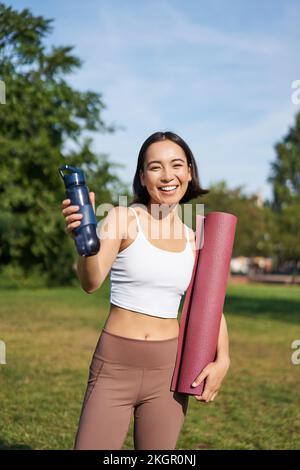 Porträt eines glücklichen asiatischen Mädchens, eine Fitnessfrau, die dir nach dem Training eine Flasche Wasser zum Trinken gibt, mit einer Gummimatte für Übungen im Park Stockfoto