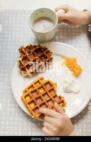 Hände eines Jungen, der eine heiße Schokoladentasse neben Waffeln in einem Teller auf dem Esstisch hielt Stockfoto