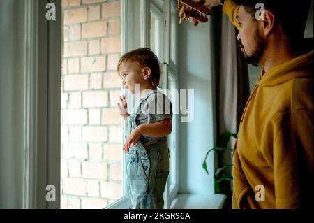 Ein süßer Junge, der durch das Fenster schaut, während der Vater zu Hause ein Flugzeugspielzeug in der Hand hält Stockfoto