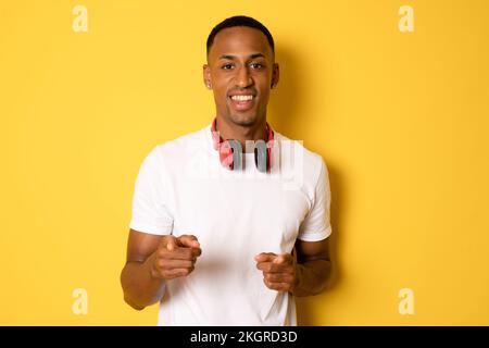 Ein junger, lächelnder afrikaner mit Kopfhörern, der auf die Kamera schaut, isoliert auf gelbem Hintergrund steht. Stockfoto
