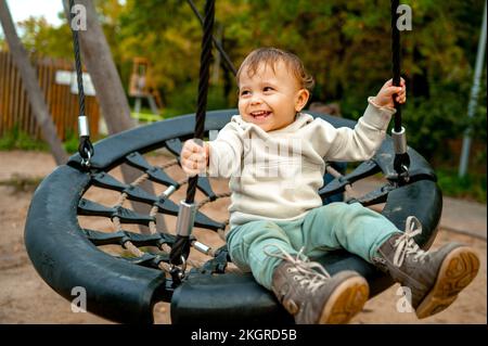 Fröhlicher süßer Junge, der auf Nestschaukel im Herbstpark schwingt Stockfoto