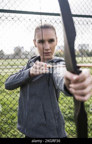 Junge Sportlerin, die vor dem Zaun Bogenschießen macht Stockfoto