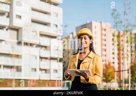 Glückliche junge Frau mit Schutzhelm, die vor Gebäuden steht Stockfoto