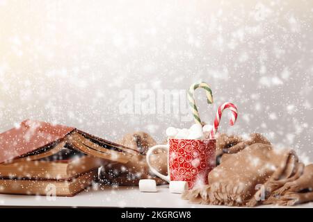 Frohe Weihnachten. Bücherstapel Tasse Marshmallow warme Decke Schal mit unscharfem Schneefall. Stockfoto