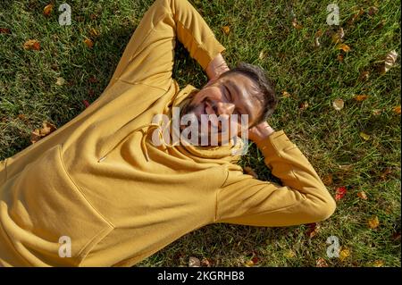 Ein fröhlicher Mann in einer gelben Kapuzenjacke, der im Park auf dem Gras liegt Stockfoto