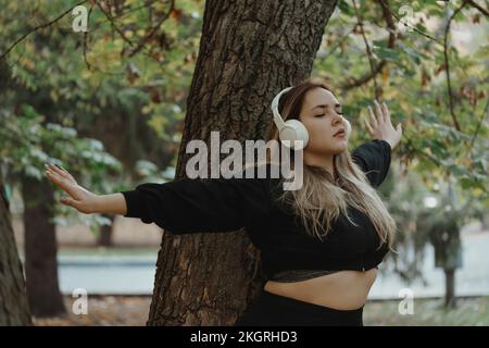 Junge Frau, die Musik über kabellose Kopfhörer hört und mit ausgestreckten Armen auf dem Baumstamm steht Stockfoto