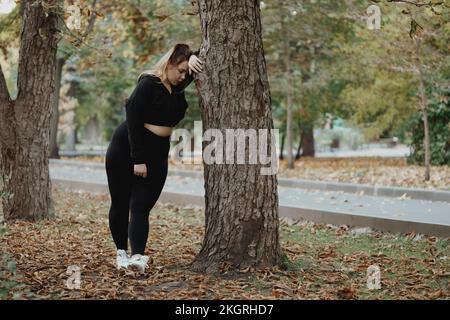 Erschöpfte Frau, die sich im Herbstpark auf den Baumstamm lehnt Stockfoto