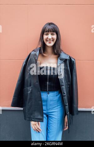 Lächelnde junge Frau mit Lederjacke vor der Wand Stockfoto