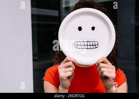 Junge Frau, die fröhliche Emoticon-Platte über dem Gesicht hält Stockfoto