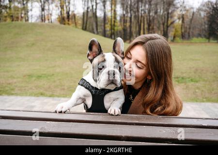 Fröhliche Frau, die französische Bulldogge auf einer Bank im Park umarmt Stockfoto