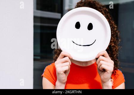 Junge Frau, die lächelnde Emoticon-Platte über dem Gesicht hält Stockfoto