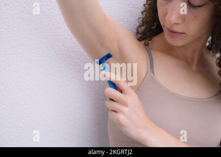 Junge Frau rasiert Achselhöhlen vor der Wand Stockfoto