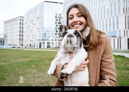 Glückliche Frau, die französische Bulldogge vor Gebäuden trägt Stockfoto