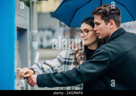 Ein junges Paar mit Schirm am Geldautomaten Stockfoto