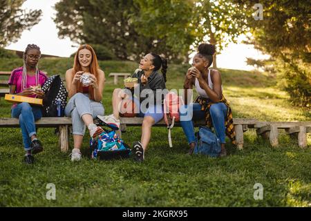 Glückliche Freunde sitzen auf einer Bank und genießen Essen im Park Stockfoto