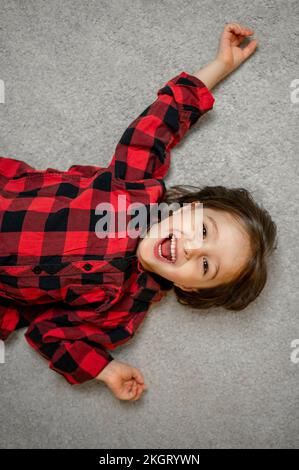 Ein fröhlicher Junge, der ein kariertes Hemd trägt und auf dem Teppich liegt Stockfoto