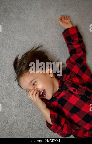 Ein glücklicher Junge mit kariertem Hemd, der auf dem Teppich lag Stockfoto